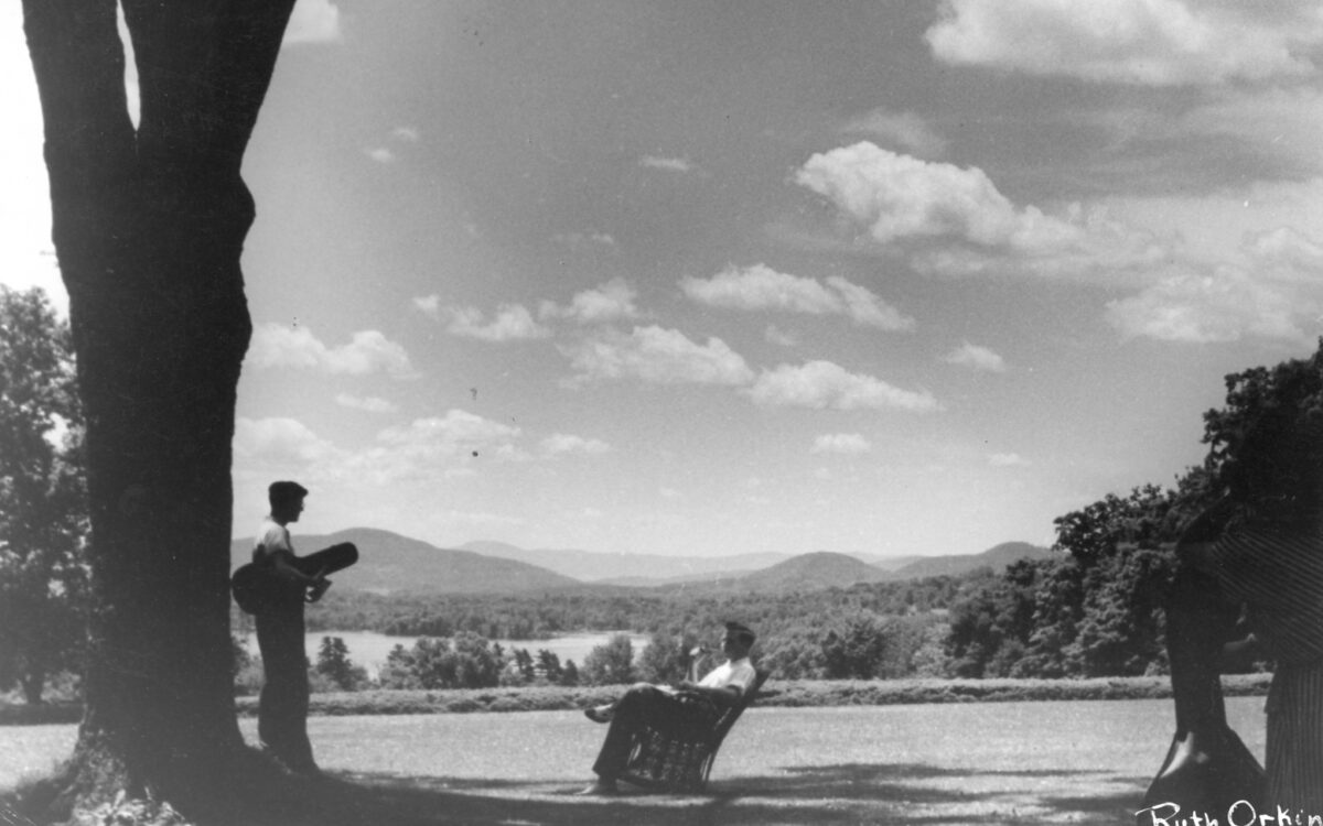 Black and white photo of Tanglewood lawn with tree in the foreground and two people conversing. One is standing under the tree holding an instrument case, the other is sitting in a chair.
