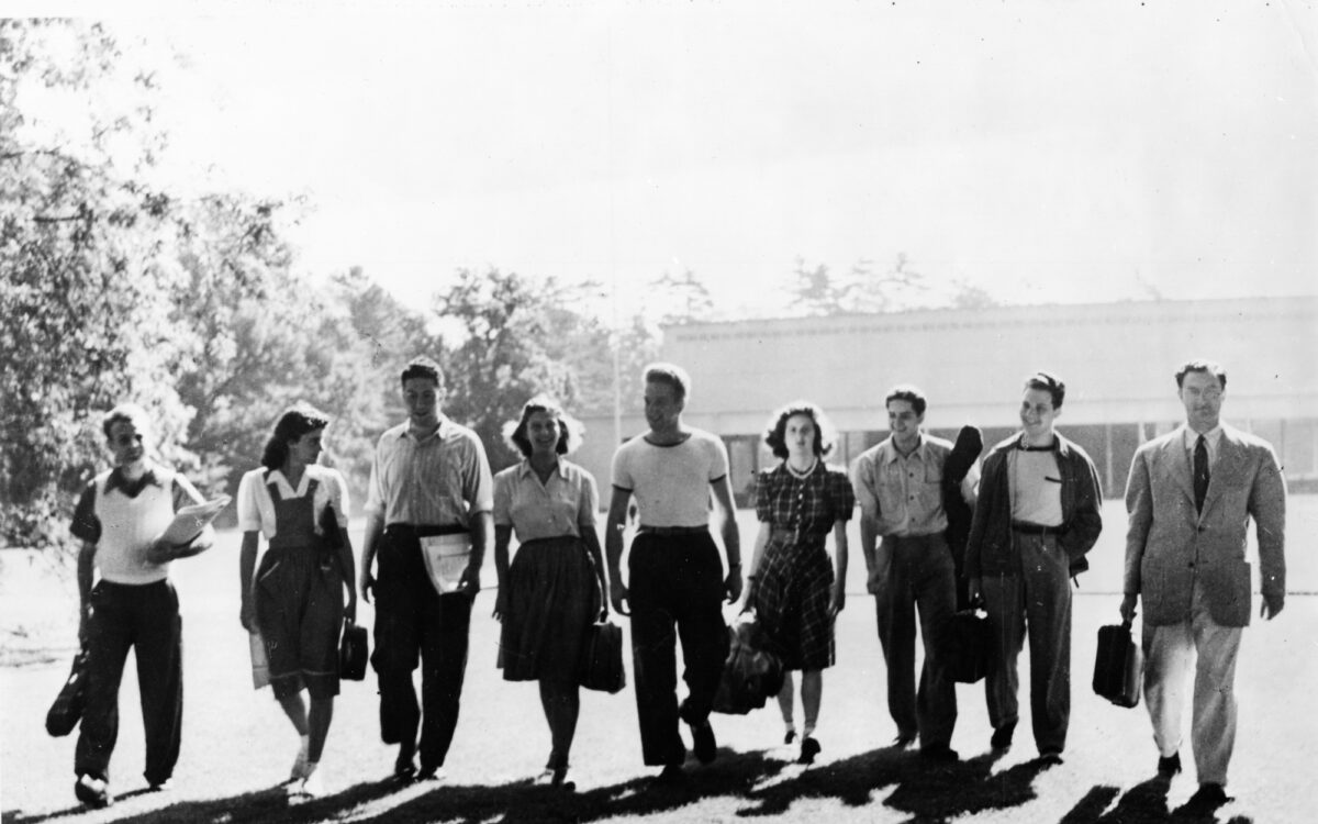 Several students holding instrument cases and wearing vintage attire walk across the Tanglewood lawn with the Music Shed in the background.