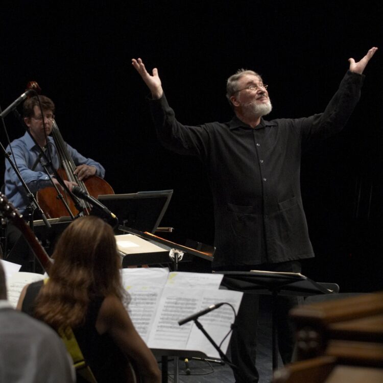 HK Gruber conducting an ensemble with his hands in the air, smiling