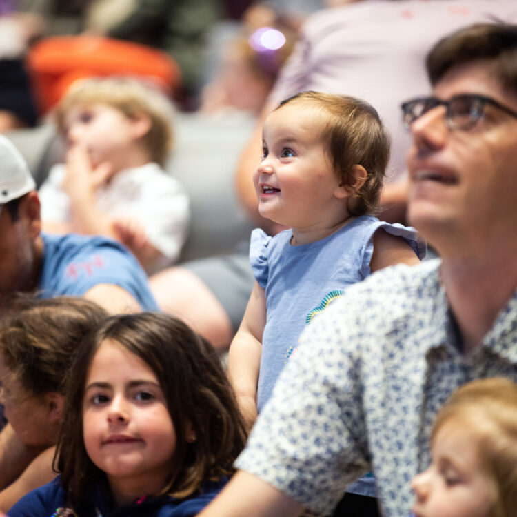 A young child smiles and looks up. They are surrounded by other children and an adult in the audience.