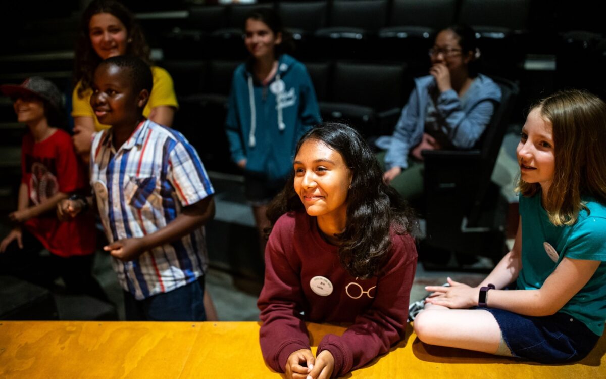Group of young students standing on the floor right in front of the stage watching performers.