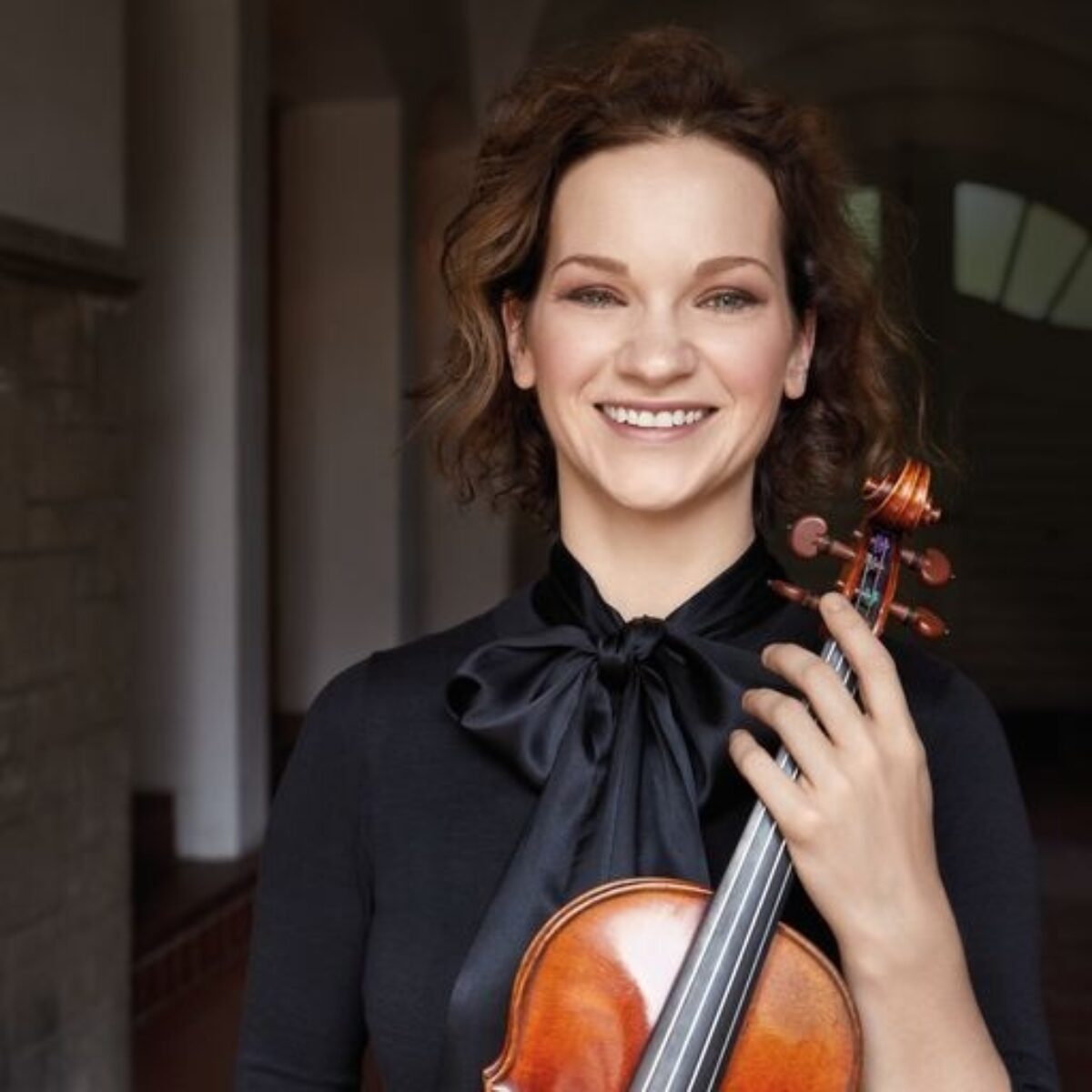 Hilary Hahn smiling directly ahead while holding a violin close to her body