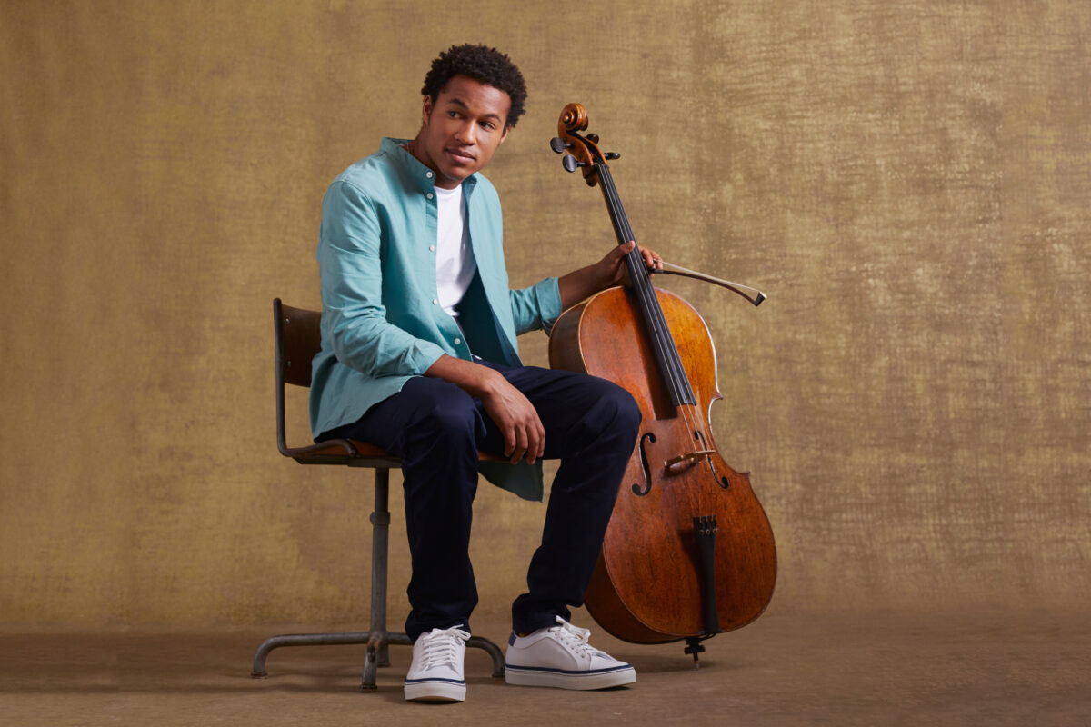 Sheku Kanneh-Mason sitting next to his cello, wearing a teal blazer