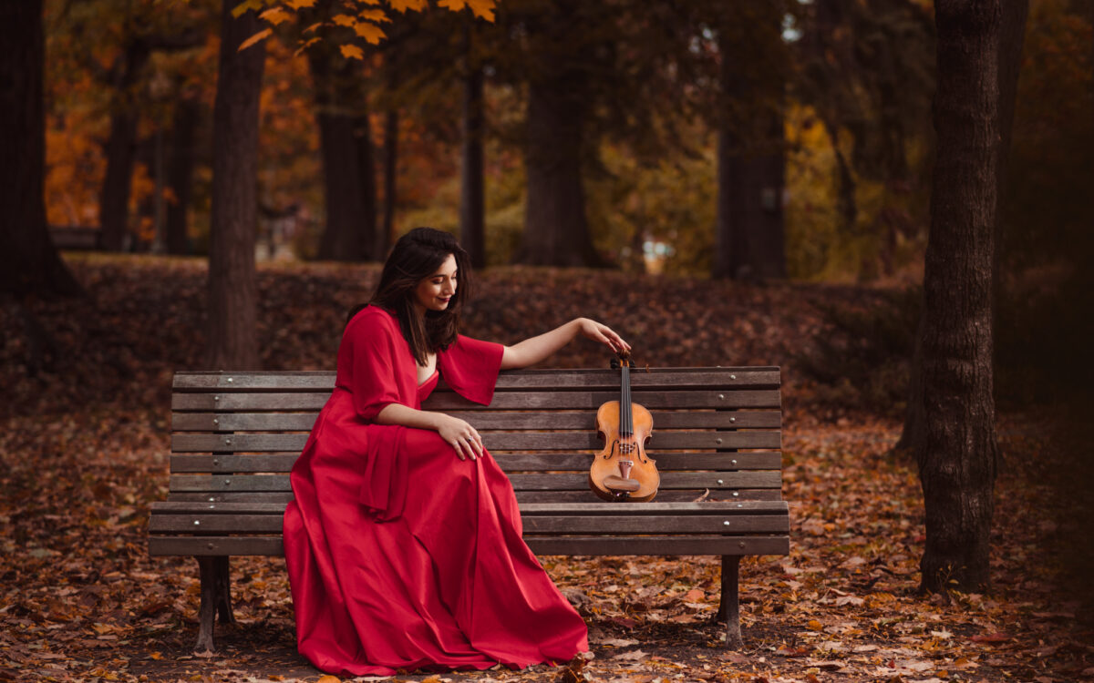 Isabella Perron sitting on a park bench wearing a long red dress, with her arm outstretched touching the neck of her violin that is next to her on the bench.