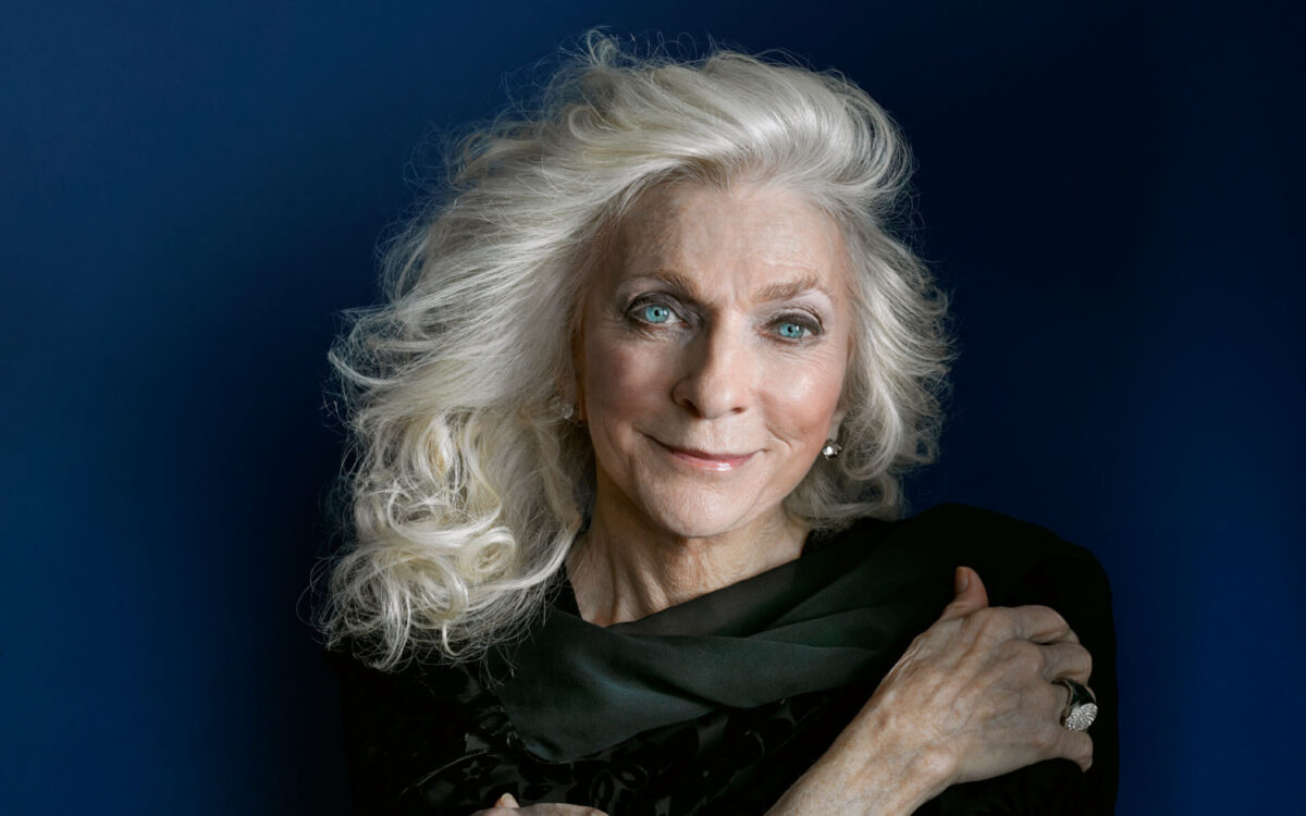 Judy Collins standing in front of a blue background with her arms crossed in front of her chest
