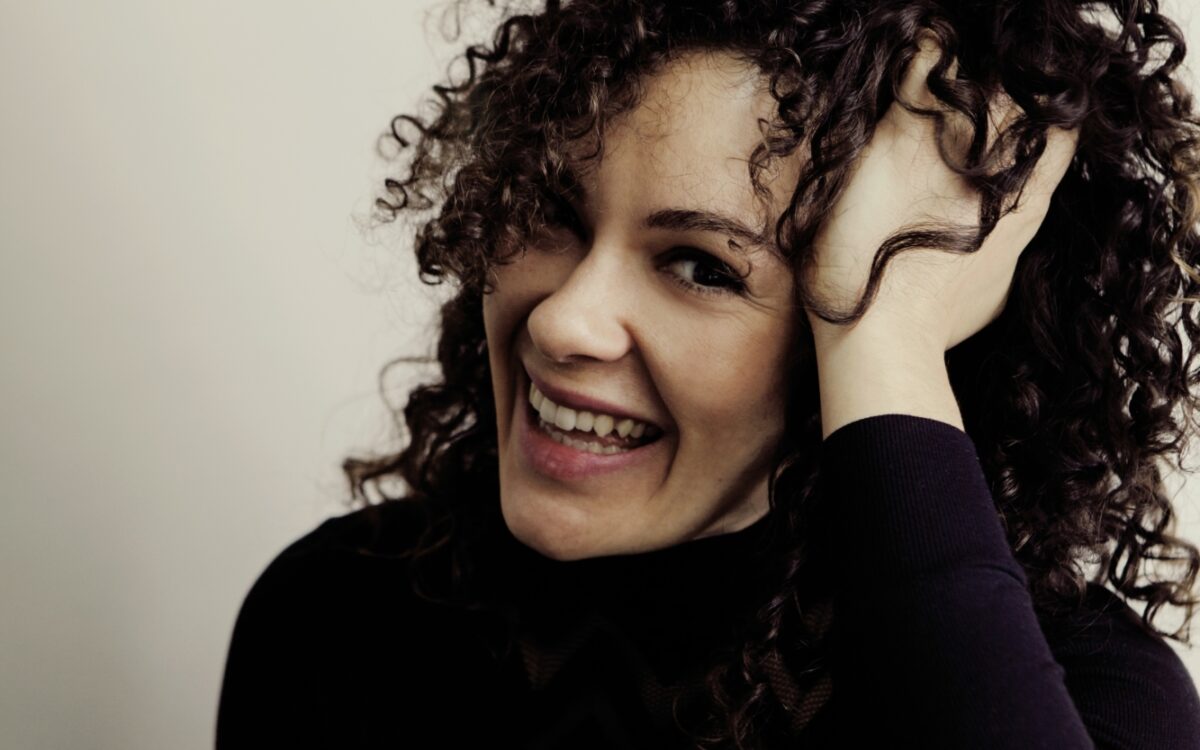 Portrait of Nora Fischer wearing a black shirt and striped pants, resting her elbow on her knee with her hand in her hair