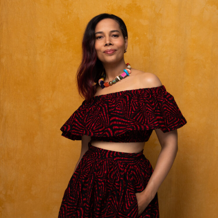 Rhiannon Giddens posing in a red patterned dress and colorful necklace with an amber backdrop