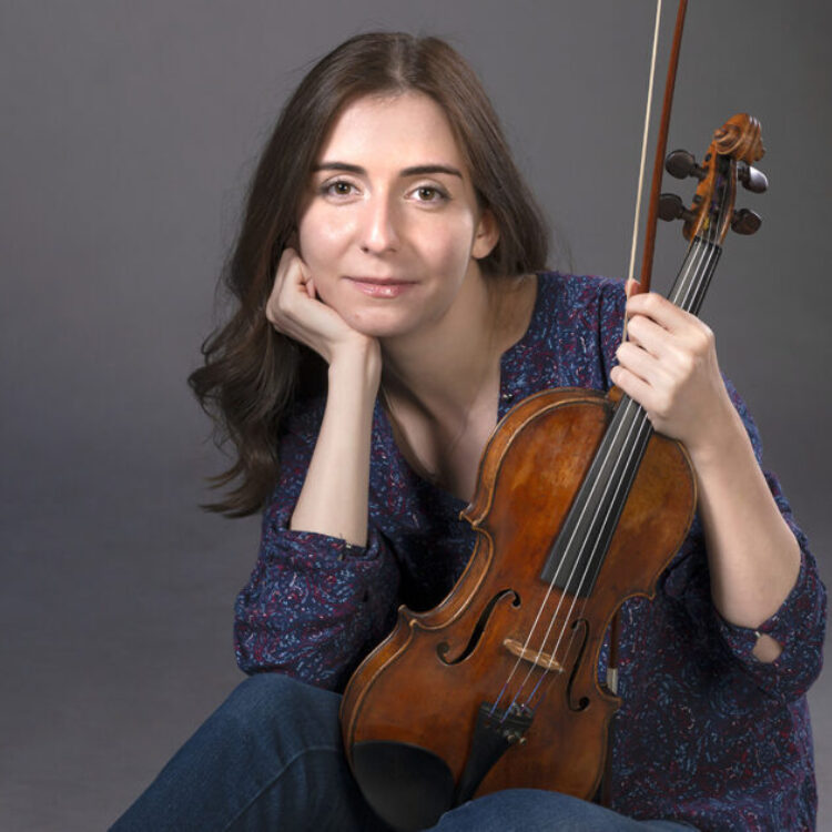 Diana Adamyan sitting down in a blue gown holding her violin and bow.