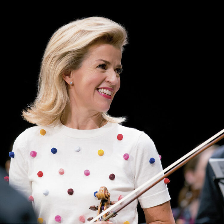 portrait of Ann Sophie Mutter holding her violin while wearing a white top with multicolored plush dots
