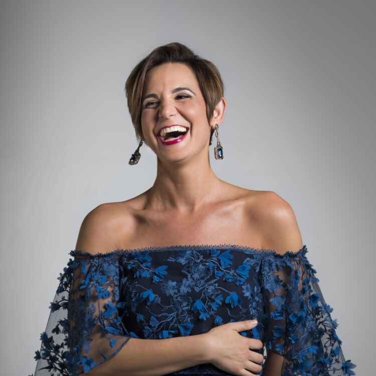 Kelley O'Connor standing in a blue dress in front of a gray background, laughing