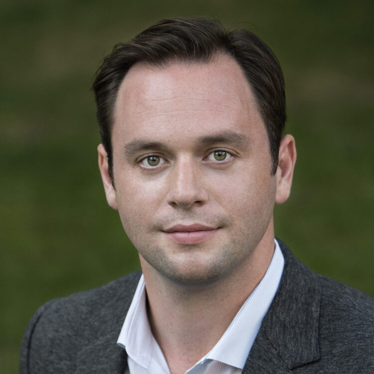 Headshot of Paul Appleby wearing a suit with grass in the background