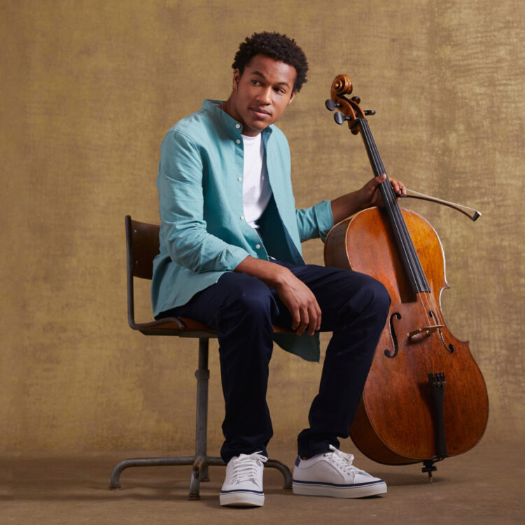 Sheku Kanneh-Mason sitting next to his cello, wearing a teal blazer