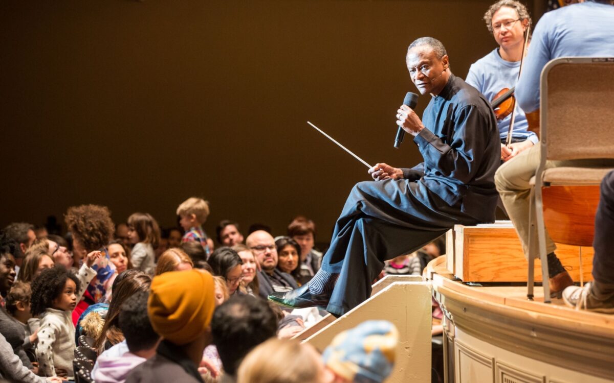 Thomas Wilkins sitting on the edge of the stage during a performance talking to an audience of children and their families.