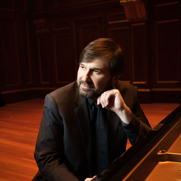 Alexander Korsantia sitting in front of a piano, smiling, with one hand on the keys and one arm resting on the top of the piano