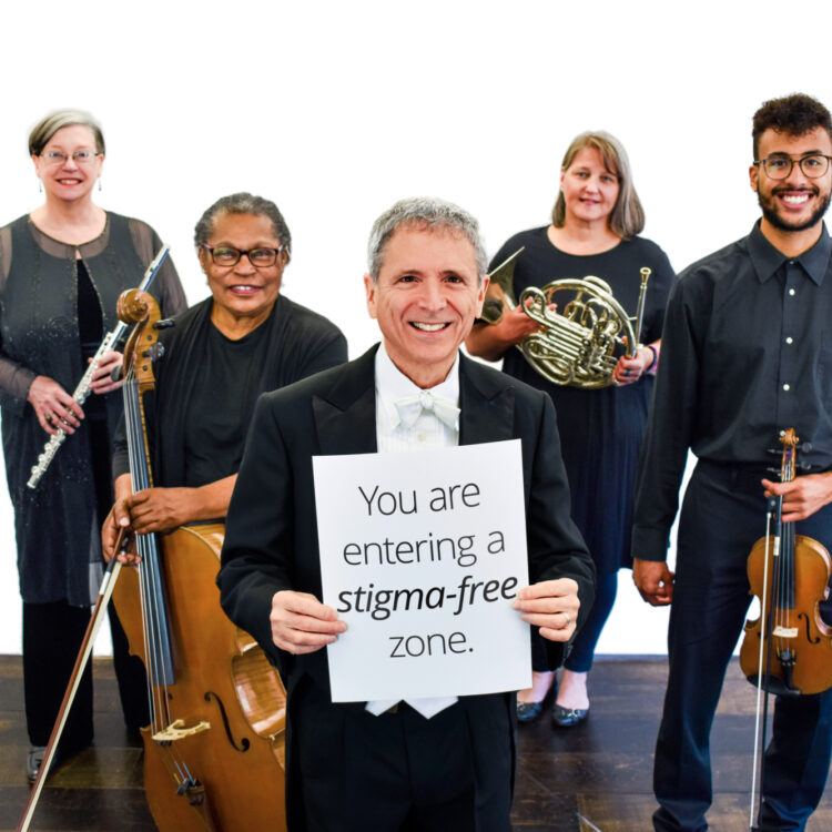 Members of the Me2 Orchestra gathered around a man in the middle who is holding a sign that reads "You are in a stigma-free zone."