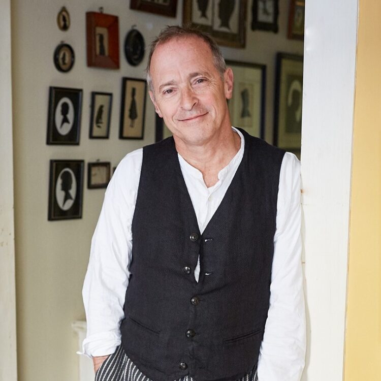 David Sedaris standing in a hallway, surrounded by framed portraits