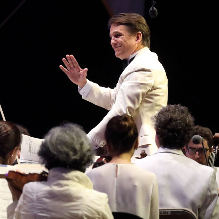 Keith Lockhart wears a white suit jacket and smiles while conducting the Boston Pops