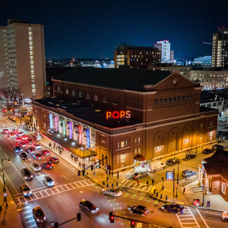 An aerial view of Symphony Hall at night, lit up by lights and a big sign that says "POPS," with lots of traffic driving by