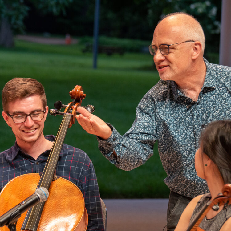A man in glasses and a blue, floral shirt gestures with his hand and speaks to music students. The young man next to him holds his cello and smiles.