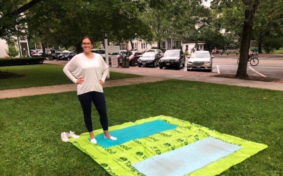A woman stands on a green tarp on some grass with two yoga mats on the tarp