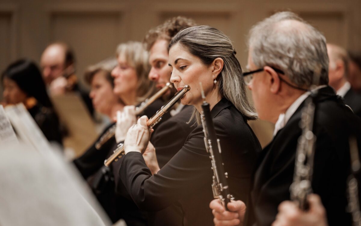 A woman with dirty-blonde hair plays the flute, with other flutists playing next to her
