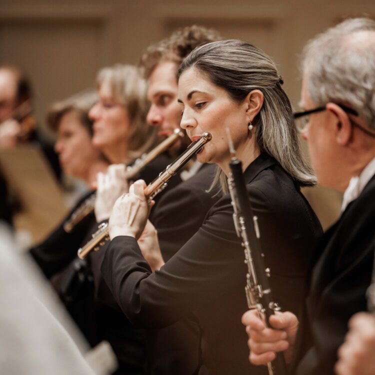 A woman with dirty-blonde hair plays the flute, with other flutists playing next to her