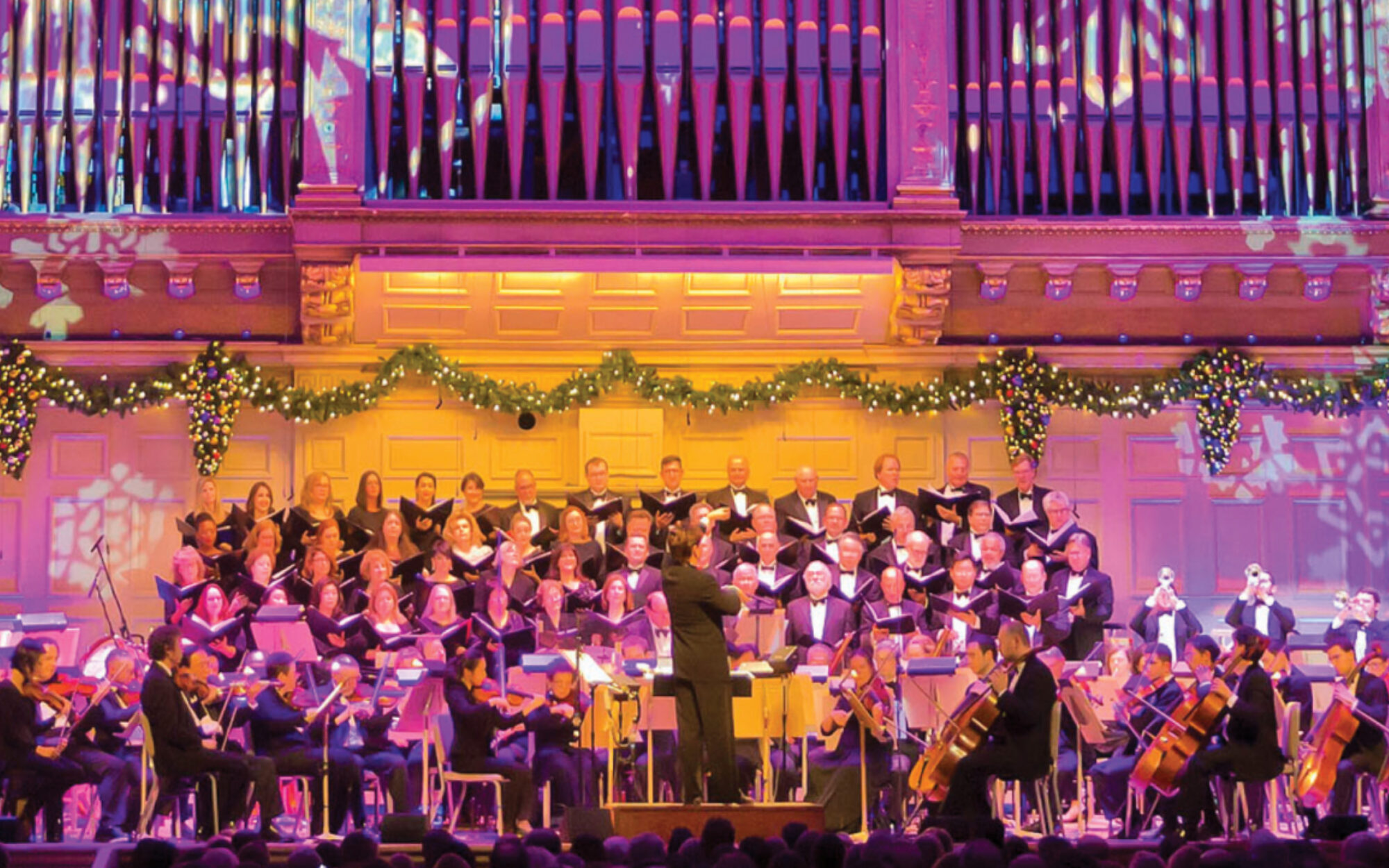The Boston Pops and Tanglewood Festival Chorus perform on the Symphony Hall stage, which is lit with a bright purple light and projections of snowflakes