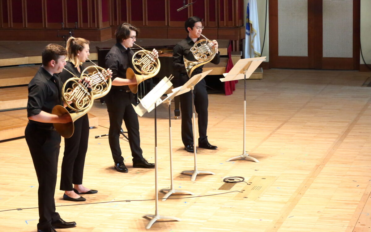 Four musicians dressed in black standing onstage playing French horns