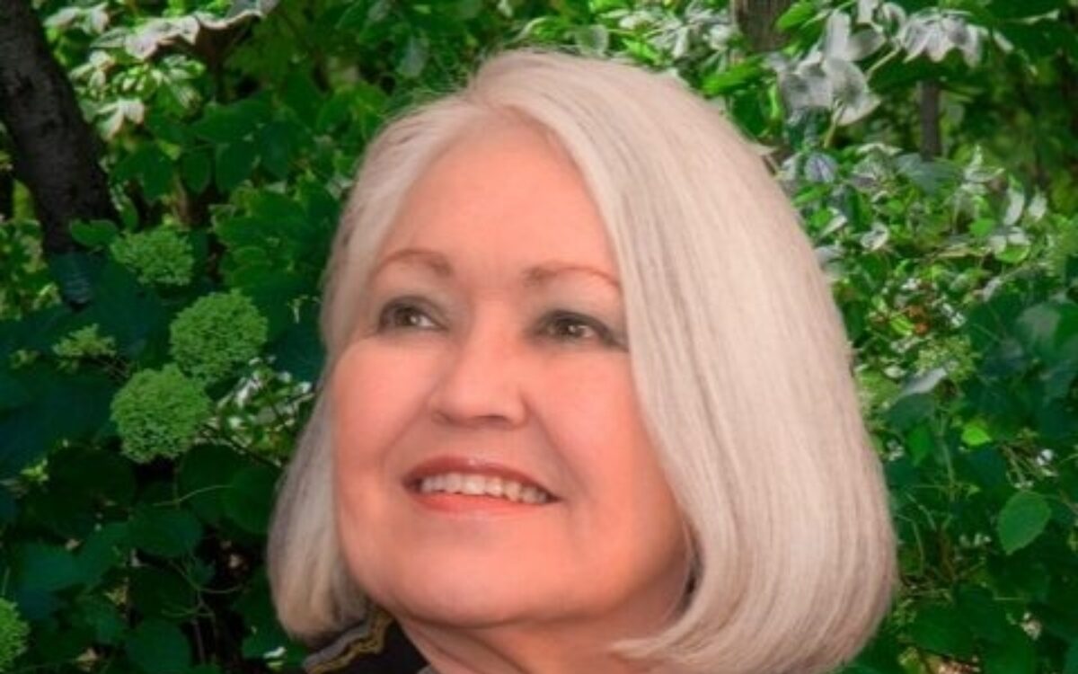Margo Garrett, wearing a black shirt with a colorlful scarf draped over her left shoulder, looking to her left and smiling