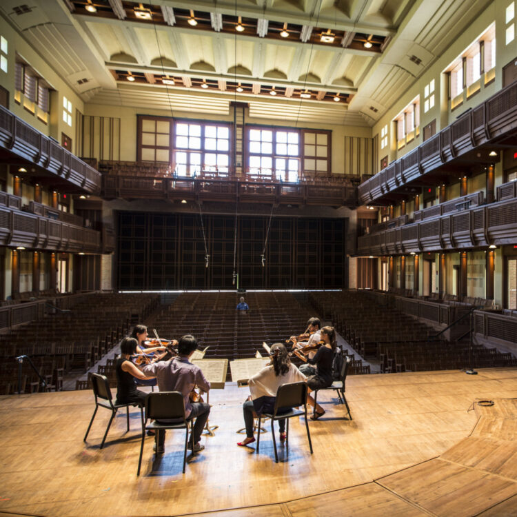 A string sextet rehearsing onstage at Ozawa Hall while a single listener watches them from the audience.