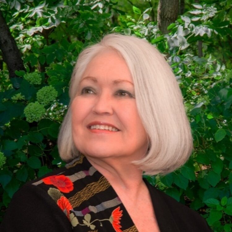 Margo Garrett, wearing a black shirt with a colorlful scarf draped over her left shoulder, looking to her left and smiling