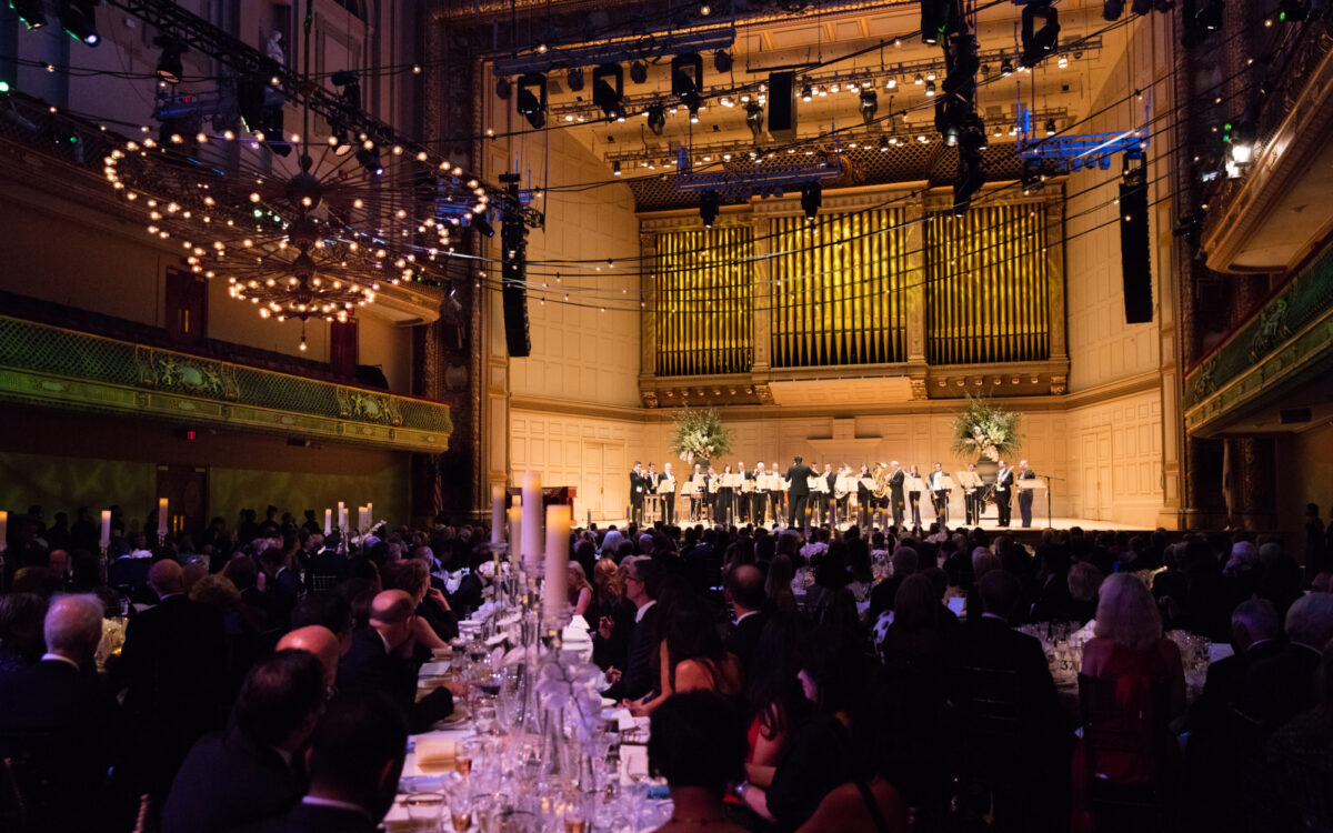 Symphony Hall decorated for a gala event with musicians performing onstage and a large chandelier illuminated.