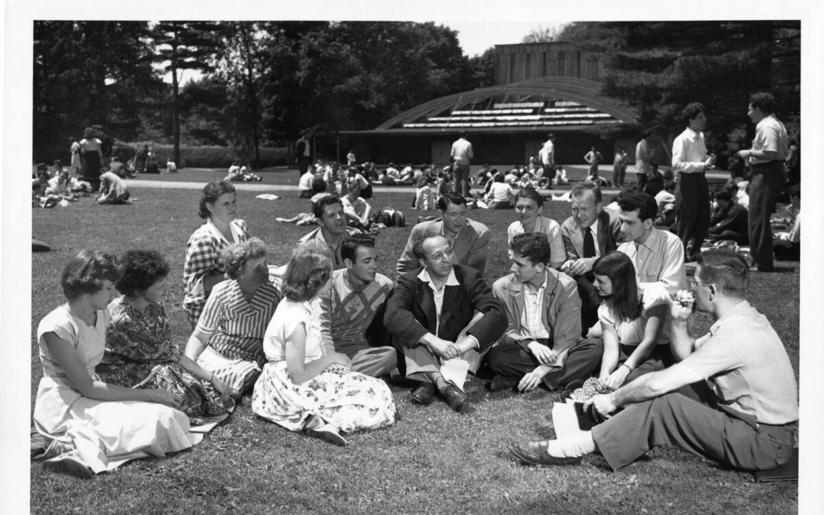 Aaron Copland sits in the middle of a group of students on the lawn in front of a building for theatre productions