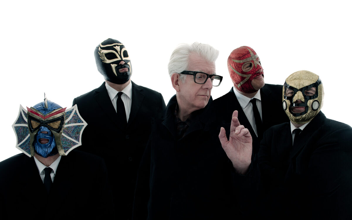 Nick Lowe wearing glasses and standing in front of four men dressed in suits and wearing face masks.