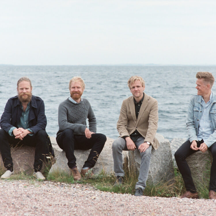 Four musicians dressed in casual clothing sitting on a rocky edge above the ocean