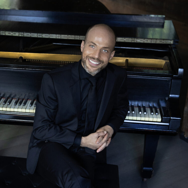 Headshot of Orion Weiss wearing a black suit and sitting in front of a piano.