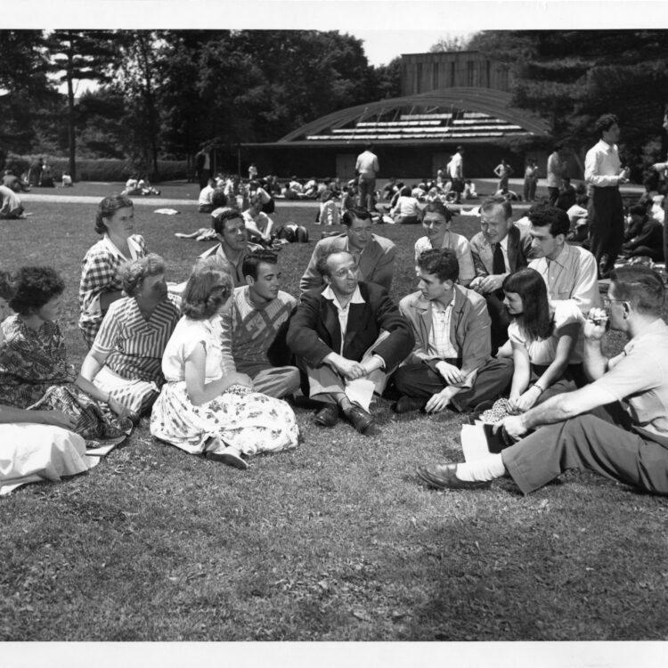 Aaron Copland sits in the middle of a group of students on the lawn in front of a building for theatre productions