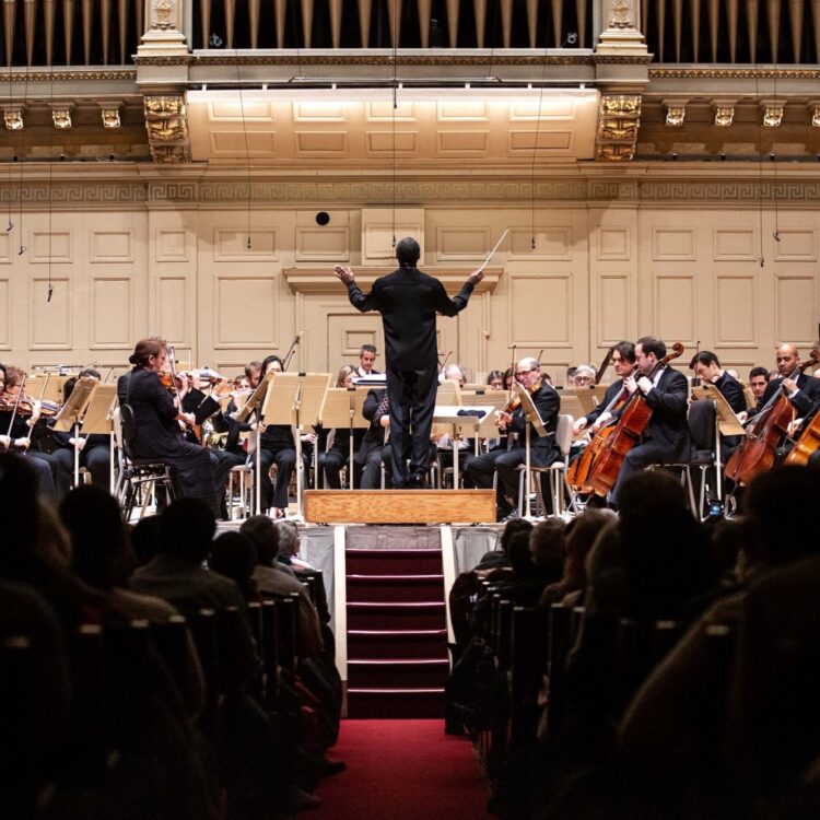 Thomas Wilkins, dressed in all black, conducts the BSO from the Symphony Hall podium as the audience watches
